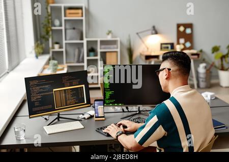 Back view of male software developer typing on keyboard with programming code on computer screen while working in office interior, copy space Stock Photo
