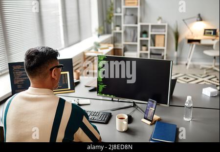 Back view of male IT developer typing on keyboard with programming code on computer screen while working in office interior, copy space Stock Photo