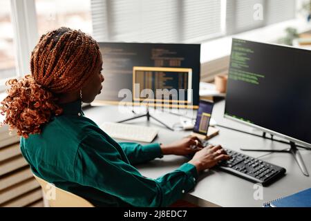 Side view of female IT developer typing on keyboard with programming code on computer screen while working in office interior, copy space Stock Photo