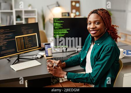 Portrait of female IT developer looking at camera and smiling against programming code on computer screen in office interior, copy space Stock Photo
