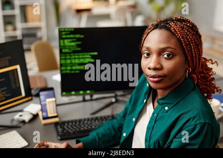 Portrait of young female IT developer looking at camera against programming code on computer screen in office interior Stock Photo