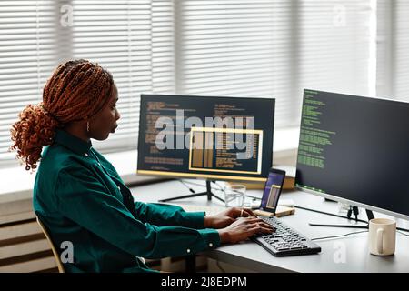 Side view of African American IT developer typing on keyboard with programming code on computer screen while working in office, copy space Stock Photo