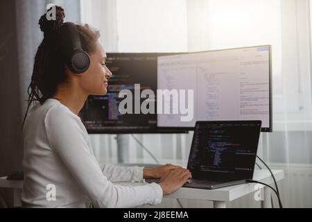 Cheerful beautiful female IT developer listening to music in headphones while coding at home Stock Photo