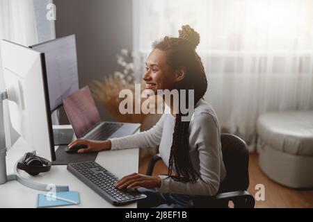 Smiled cheerful female web expert sitting at desk in living room coding on computer Stock Photo
