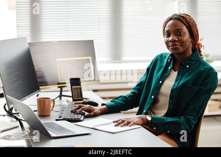Warm toned portrait of black young woman as IT developer or QA engineer smiling at camera while sitting at workplace with computers, copy space Stock Photo