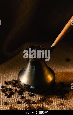 An old copper cezve stands on a dark background. Coffee beans are scattered on burlap. Stock Photo