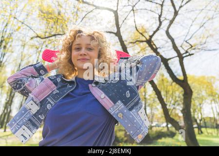 Joyful curly-haired european woman in her 20s wearing a spring patterned jacket, standing in a blooming park, posing with a skateboard. . High quality photo Stock Photo