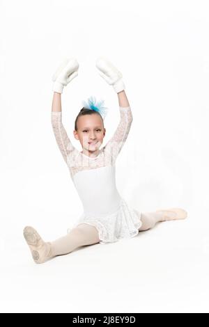 Smiling girl ballet dancer nine-year-old in white ballet tutu skirt, blue bow in hair and white mittens ballet stretching on white floor. Studio shot. Stock Photo
