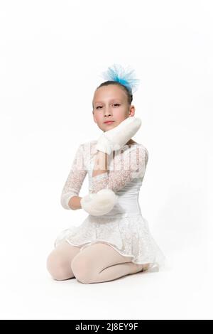 Girl ballet dancer nine-year-old in white ballet tutu skirt, blue bow in hair and white mittens sitting on white floor. Studio shot Stock Photo
