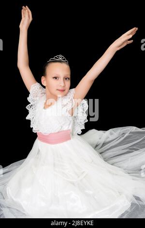 Girl ballerina in white long dress sitting on black background and poses elegantly with arms raised. Caucasian ballet dancer nine-year-old Stock Photo
