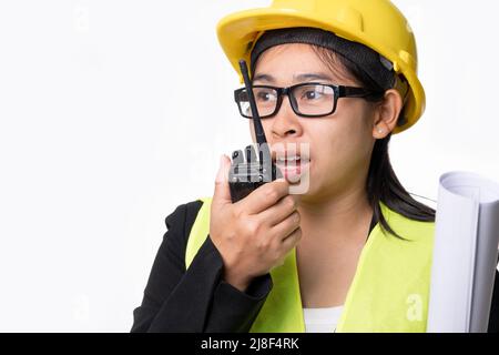 Female civil engineer in a helmet holding construction plans and using walkie-talkie and talk to other staff on a white background in studio. Stock Photo