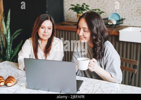 Adult smiling brunette middle aged women sisters friends having breakfast and using laptop in kitchen at home Stock Photo