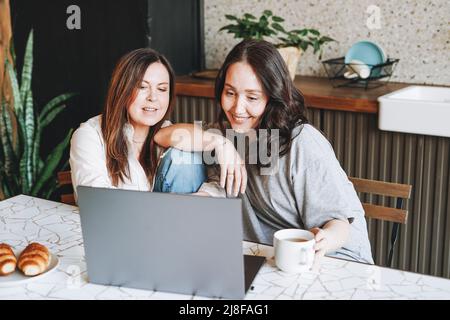 Adult smiling brunette middle aged women sisters friends having breakfast and using laptop in kitchen at home Stock Photo