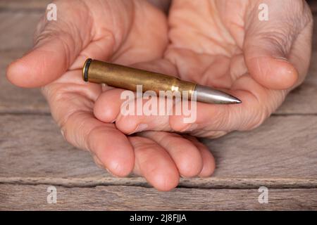 The hands of an old woman hold a military bullet on a wooden background, the war in Ukraine, the protection of civilians, a woman military Stock Photo