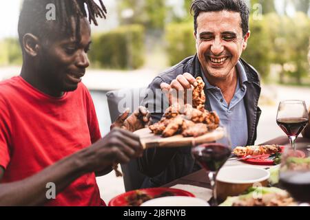 Happi multi-ethnic friends sitting at farmhouse table - an African guy hands the grilled chicken wings to a mid aged man - conviviality and friendship Stock Photo
