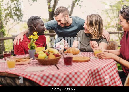 Multiethnic group of people gather on farm in the morning for breakfast picnic - friends sitting at an outdoor table drinking fruit juices and eating Stock Photo