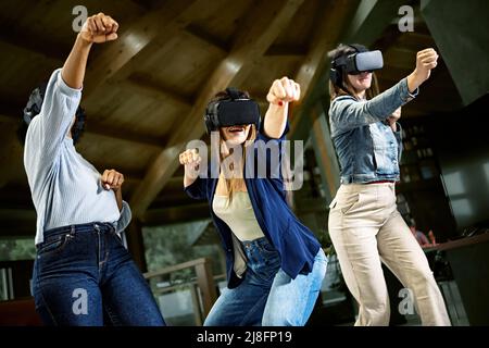Group of young women using virtual reality headset - Girlfriends playing ninja fight video games in meta verse space with futuristic goggles having fu Stock Photo
