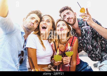 Happy drunk friends taking a selfie making faces holding alcoholic cocktails sitting on a bench outdoors in the summer - friendship love in happy hour Stock Photo