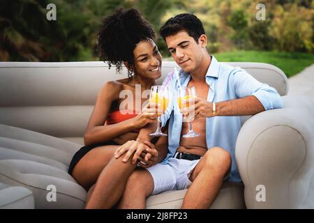 Biracial couple of gen z young newlyweds people sitting on the luxury resort patio talking carefree and drinking fruit juice mocktails - healthy and f Stock Photo