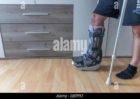 European man after Achilles tendon rupture operation is back home with moon boot special physiotherapy shoe and crutches for recovery at home against Stock Photo