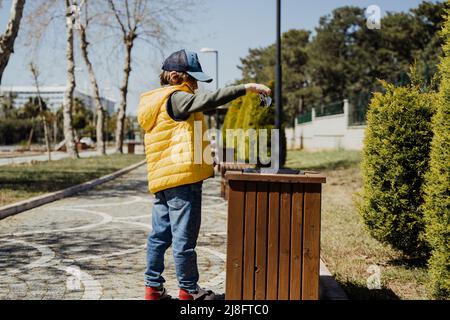 Schoolboy kid throwing the trash into dumpster. Boy using recycling bin to throw away the litter. Caucasian child recycles the junk into the trash-can Stock Photo