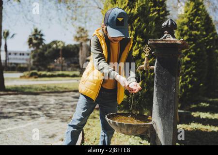 Kid schoolboy in casual clothing washing his hands in the street old fashioned drinking water fountain. Boy in blue jeans and yellow vest playing with Stock Photo