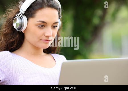 Concentrated woman with wireless headphones watching media on laptop in a park Stock Photo