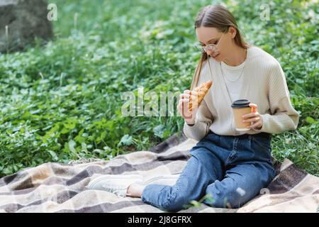woman in glasses holding paper cup and croissant while sitting on blanket during picnic Stock Photo