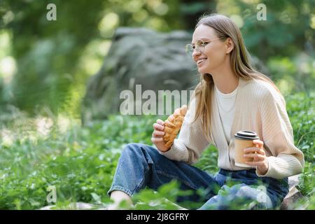 happy woman in glasses holding paper cup and croissant while sitting on grass during picnic Stock Photo