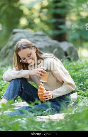 joyful woman in glasses holding paper cup and croissant during picnic Stock Photo