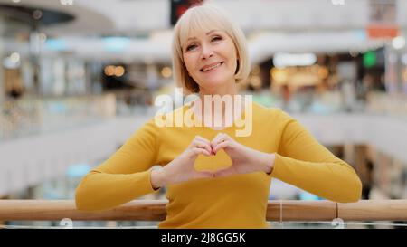 Mature happy caucasian woman standing in public place posing sincerely smiling looking at camera making heart shape with fingers sign generosity and Stock Photo
