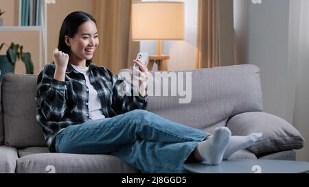 Asian girl woman sitting on sofa relaxing at home on couch playing mobile game wins getting new job offer message online betting reads good news phone Stock Photo