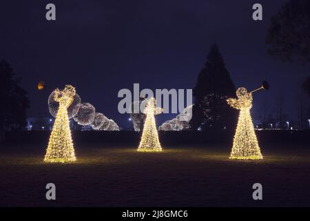 Three Christmas angels from luminous garlands illuminate the park walking area against backdrop of round luminous trees at night. Festive decor Stock Photo