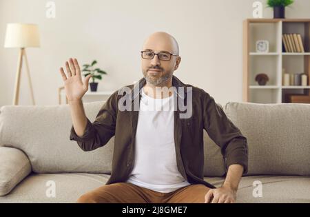 Video call portrait of mature man sitting on sofa at home and waving hand at camera Stock Photo