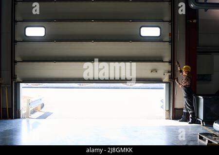 Young female worker of factory or hangar in coveralls and protective helmet opening shutter roller door at the beginning of working day Stock Photo