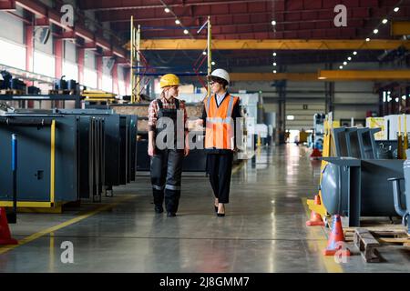 Two young female factory workers in protective helmets having discussion of working points on the move along large workshop Stock Photo