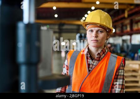 Young female worker of modern factory in orange jacket and protective helmet looking at camera in large workshop or warehouse Stock Photo