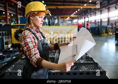 Side view of young female engineer in workwear holding unrolled blueprint with sketch of construction or machine part Stock Photo
