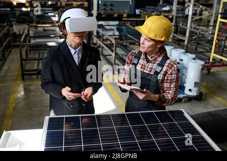 Two young engineers of factory discussing virtual model of new product while one of them using tablet and talking to woman in vr headset Stock Photo