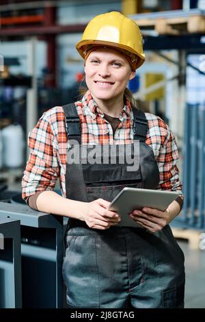 Young successful female engineer in coveralls and hardhat holding tablet while standing in front of camera against interior of warehouse Stock Photo