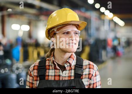 Happy young female engineer in hardhat and coveralls standing in workshop or warehouse of factory with industrial equipment Stock Photo