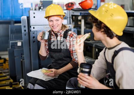 Young smiling female engineer with sandwich and cup of tea talking to colleague at lunch break while both sitting on staircase Stock Photo