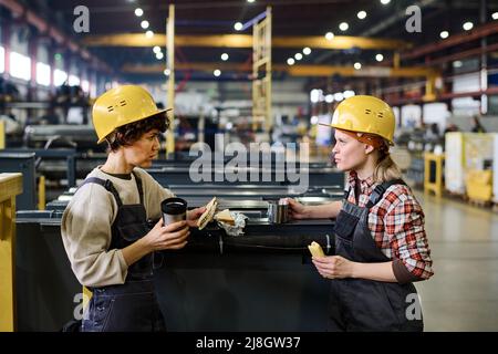 Two young engineers or workers of warehouse having sandwiches and hot tea at lunch break and discussing latest news or working points Stock Photo
