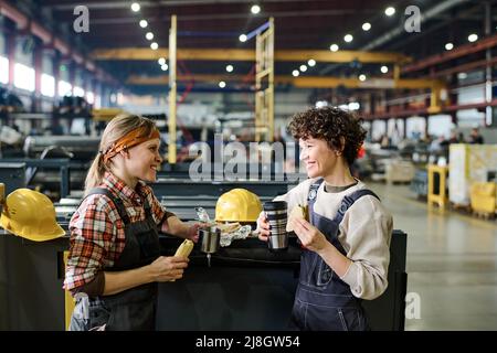 Two happy female engineers in coveralls looking at one another during talk by lunch while having sandwiches and hot tea in workshop Stock Photo