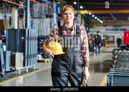 Happy young female engineer in coveralls and checkered shirt holding protective helmet in hand while standing in front of camera Stock Photo