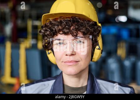 Face of young engineer in protective helmet, eyeglasses and headphones looking at camera while standing against industrial equipment Stock Photo