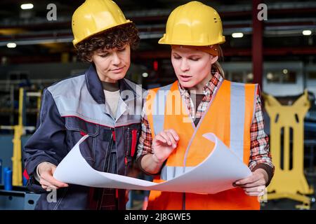 Two young female engineers discussing sketch on blueprint at working meeting at factory while one of them commenting its details Stock Photo