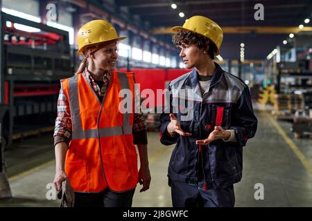 Two young female workers of modern factory discussing working points while moving down large workshop or distribution warehouse Stock Photo
