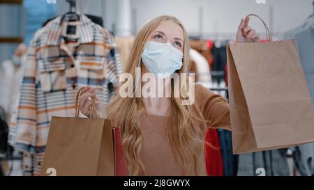 Rich happy blonde woman shopper wearing medical face protective mask girl customer client in clothing boutique with shopping bags dancing enjoying Stock Photo