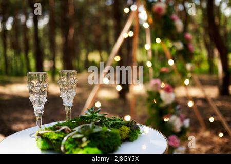 Wine glasses, wedding rings bedding of grass and moss on white table next  to bohemian tipi arch decorated in boho style with flowers and candles and  Stock Photo - Alamy